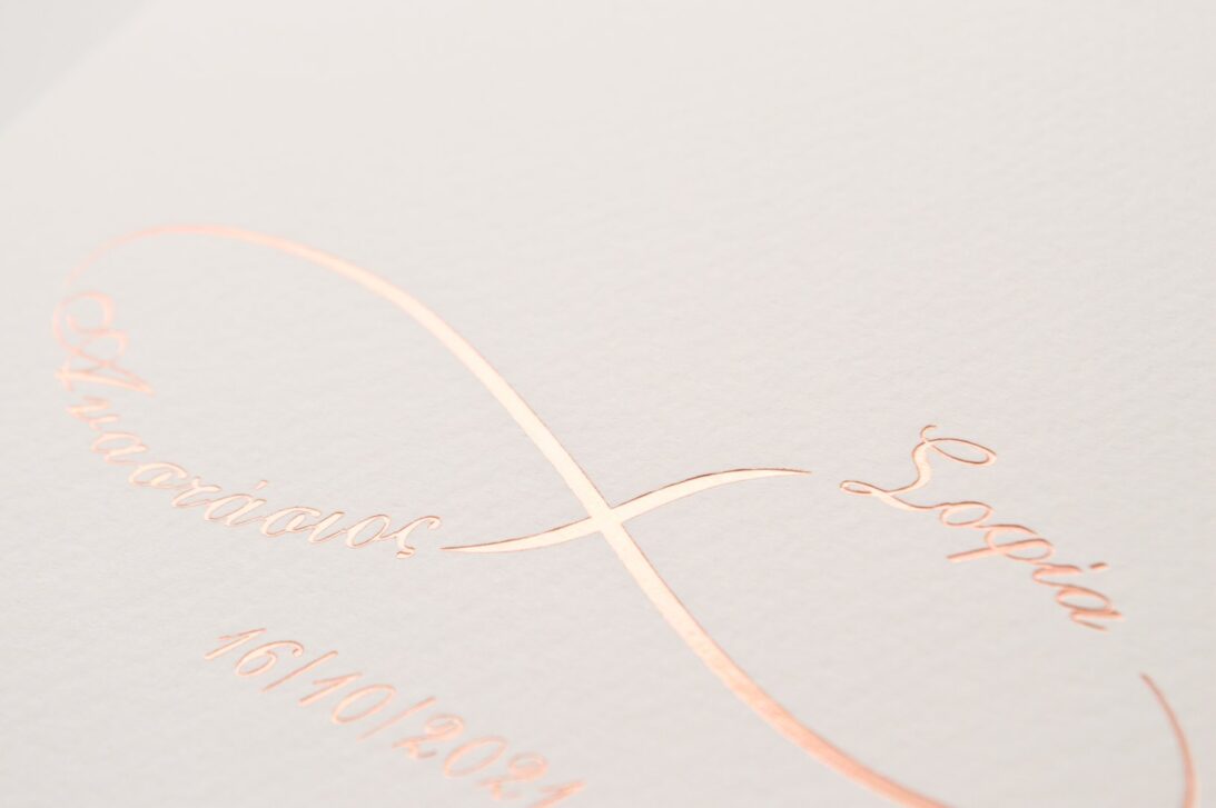 Λεπτομέρεια Προσκλητηρίου Γάμου: Φάκελος τραβηχτός διάστασης 14,5x20,5 εκατ. γκοφρέ (ανάγλυφο) μπιμπικωτό λευκό 250γραμ. και μονογράμματα σε θερμοτυπία ροζ χρυσό με θέμα άπειρο και ονόματα ζευγαριού και Κάρτα σε χαρτί (ανάγλυφο) μπιμπικωτό λευκό 250 γραμ. με εκτύπωση μελάνι γκρι και ροζ χρυσό και θέμα άπειρο και ονόματα ζευγαριού_Κωδικός 0027
