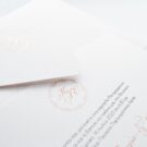 Λεπτομέρεια Προσκλητηρίου Γάμου και Βάπτισης: Φάκελος διάστασης 16x22 εκατ. σε χαρτί γκοφρέ μπιμπικωτό λευκό 150γραμ. με μοτίβο στεφανάκι και μονογράμματα σε εκτύπωση ροζ χρυσοτυπία και Κάρτα σε χαρτί γκοφρέ μπιμπικωτό λευκό 250 γραμ. με εκτύπωση μοτίβου στεφανάκι και μονογραμμάτων με μελάνι βρώμικο ροζ και κείμενο με εκτύπωση σε μελάνι γκρι και βρώμικο ροζ_Κωδικός_ΓΒΝΣ 2002