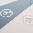 Λεπτομέρεια Προσκλητηρίου Γάμου και Βάπτισης: Φάκελος διάστασης 16x22 εκατ. σε χαρτί οικολογικό με άγρια υφή βρώμικο γαλάζιο 160γραμ. με μοτίβο στεφανάκι και μονογράμματα σε εκτύπωση ασημοτυπία γυαλιστερή και Κάρτα σε χαρτί οικολογικό βελούδο υπόλευκο 250 γραμ. με εκτύπωση μοτίβου στεφανάκι και μονογραμμάτων με μελάνι βρώμικο γαλάζιο και κείμενο με εκτύπωση σε μελάνι γκρι και βρώμικο γαλάζιο_Κωδικός_ΓΒΝΣ 2003